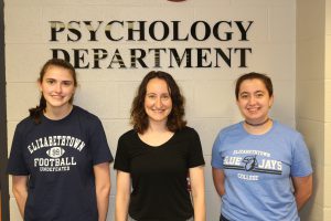 Two Etown students and their prfessor smiling at camera in front of Psychology department sign.