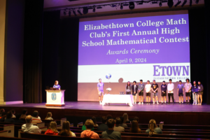 High school students stand on stage at Leffler Chapel in front of a screen that reads "Elizabethtown College Math Club's First Annual High School Mathematical Contest Awards Ceremony." Audience members watch.
