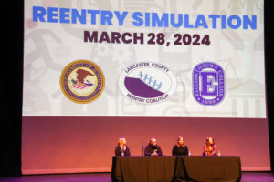 Four adults sit at a table on a stage, behind them is a slideshow that reads "Reentry Simulation." 
