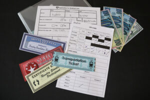 Simulation documents on a table, including a transportation ticket, a certificate of birth, money, a social security card, and a schedule of events. 